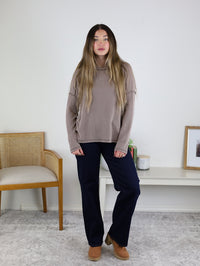 A woman with long hair wears a taupe ribbed knit Finley Sweater, dark pants, and brown shoes, standing in a room with a chair, white table, books, framed art, and a light rug.