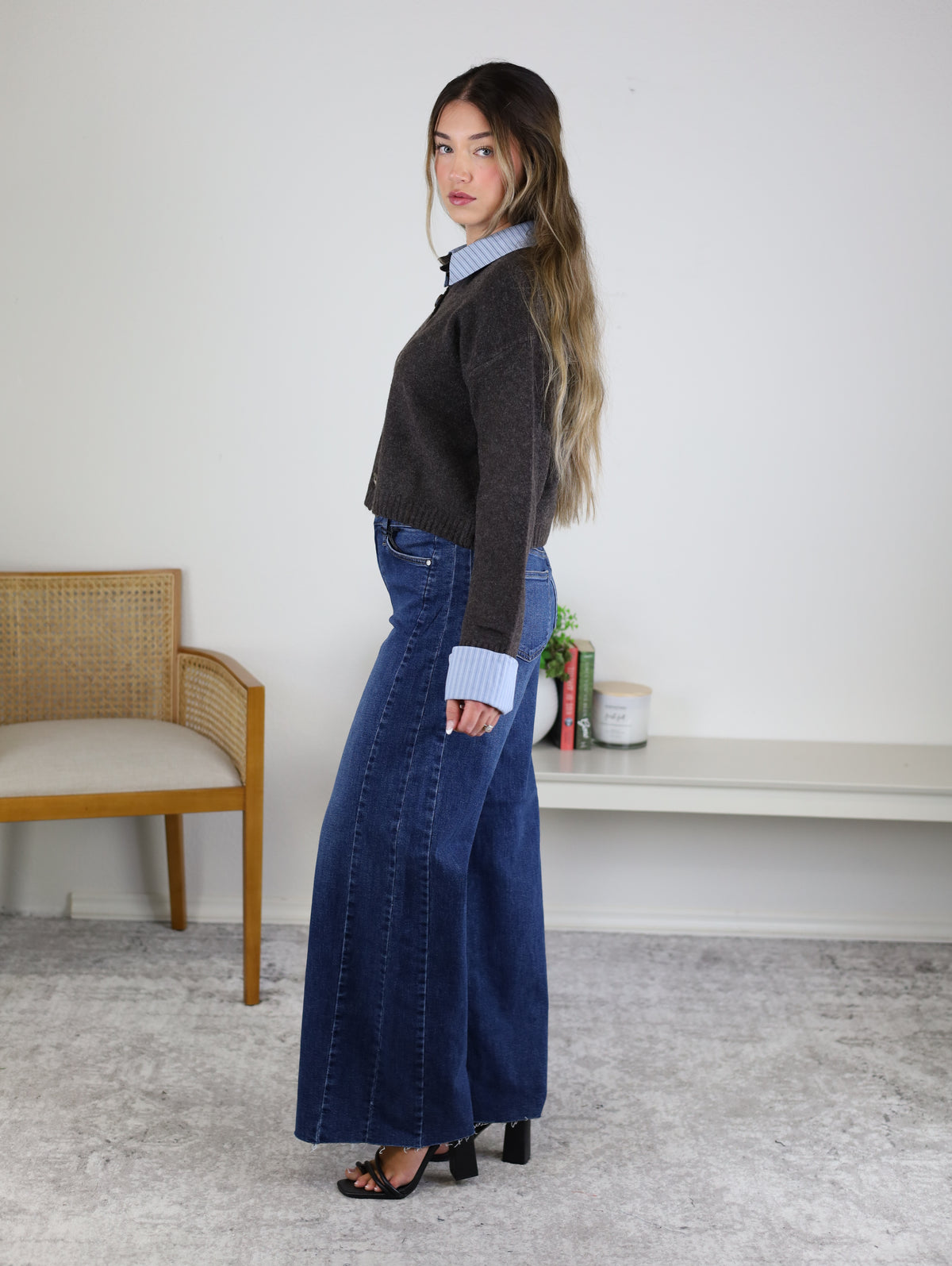 A woman stands sideways in a room, wearing the Mani Stripe Trim Button Down Cardigan over a blue shirt, wide-leg jeans, and black heels. There’s a chair, a shelf, and a small plant in the background.