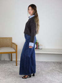A woman stands sideways in a room, wearing the Mani Stripe Trim Button Down Cardigan over a blue shirt, wide-leg jeans, and black heels. There’s a chair, a shelf, and a small plant in the background.