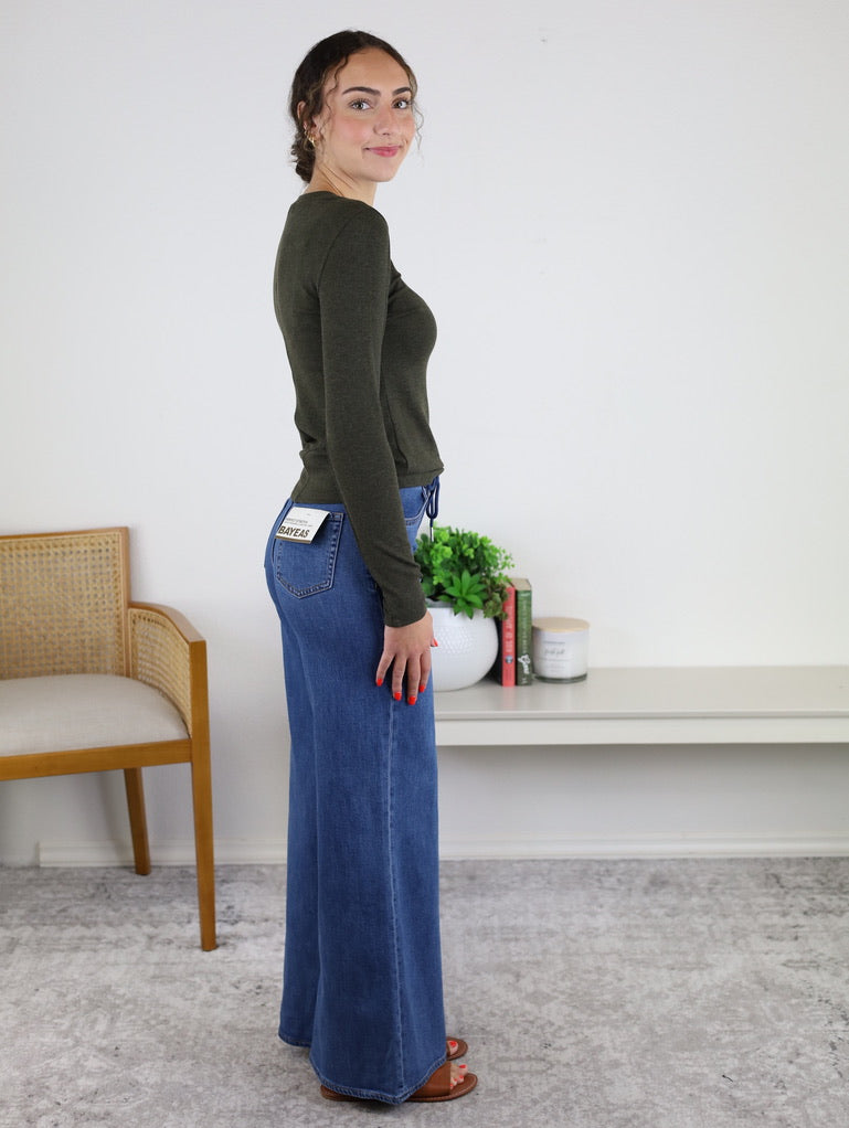 A woman stands sideways in a minimal living space with books, a plant, and a chair in the background, wearing the fitted Josette Ribbed Classic Crewneck in dark green and high-waisted wide-leg blue jeans.
