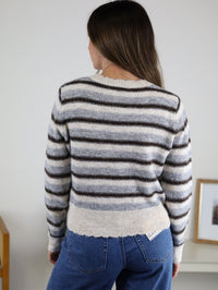 A person wearing the Lark Lace Trim Striped Knit Cardigan with Candy Buttons, a cropped, grey, white, and brown striped knit sweater with a lace-trimmed hem.