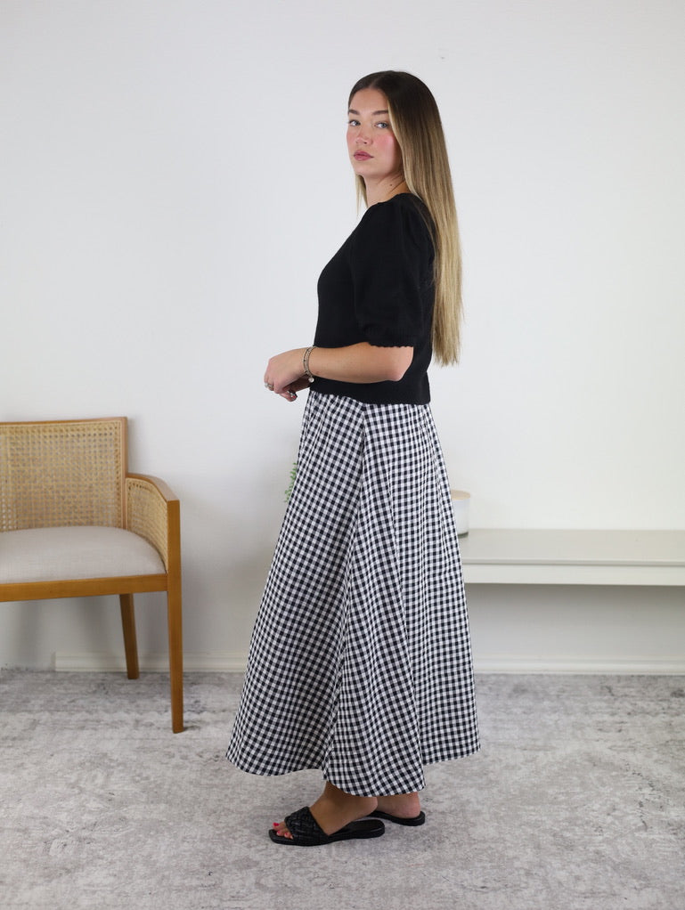 A woman stands sideways in a room, wearing the Monica Short Sleeve Cardigan with puff sleeves, paired with a black and white gingham skirt and black sandals. Her long hair is down. In the background, there is a wooden chair and a bench against a white wall.