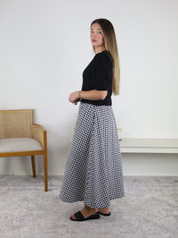 A woman stands sideways in a room, wearing the Monica Short Sleeve Cardigan with puff sleeves, paired with a black and white gingham skirt and black sandals. Her long hair is down. In the background, there is a wooden chair and a bench against a white wall.