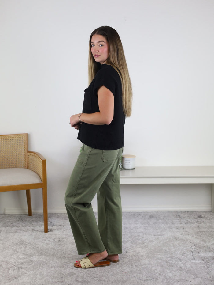 A woman with long, straight hair stands sideways indoors near a wooden chair and a white bench against a plain white wall. She is wearing a black top and the Josie Stretch Twill Utility Barrel jeans in olive green, featuring wide legs and utility pockets, styled with beige sandals.