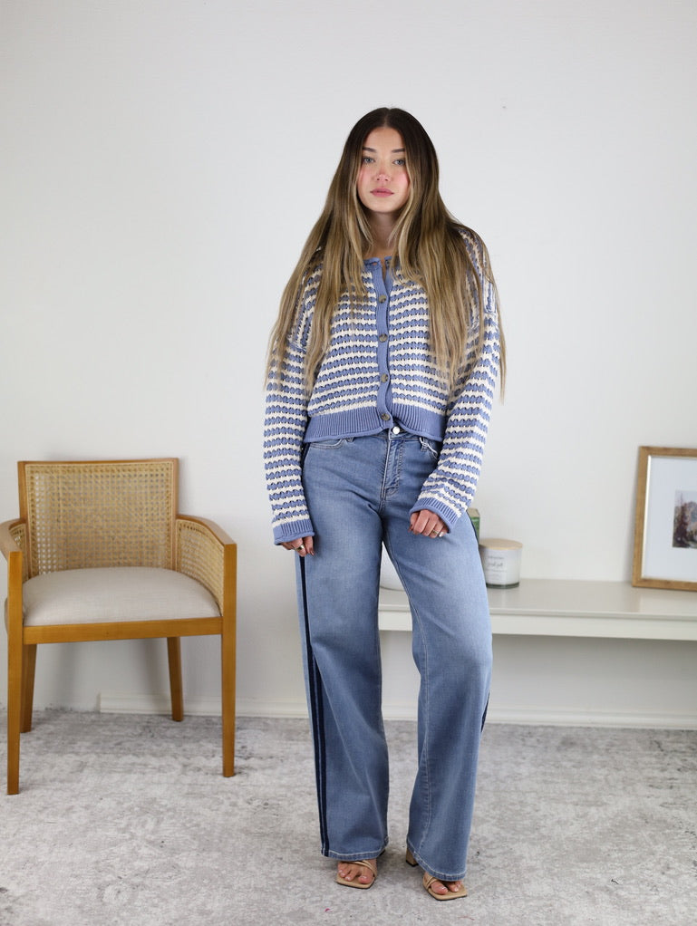 A woman with long hair wears the Bellamy Textured Striped Crochet Cardigan and wide-leg jeans, standing indoors next to a wooden chair and a white shelf with décor against a plain wall.