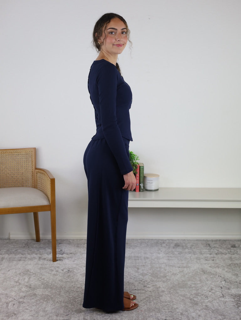A woman stands sideways indoors, wearing the Statler Lounge Set in navy, which features a long-sleeve henley top and matching wide-leg pants paired with brown sandals. In the background, there is a chair, a white shelf, several books, and some candles.