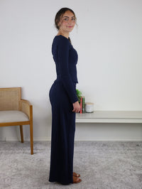 A woman stands sideways indoors, wearing the Statler Lounge Set in navy, which features a long-sleeve henley top and matching wide-leg pants paired with brown sandals. In the background, there is a chair, a white shelf, several books, and some candles.