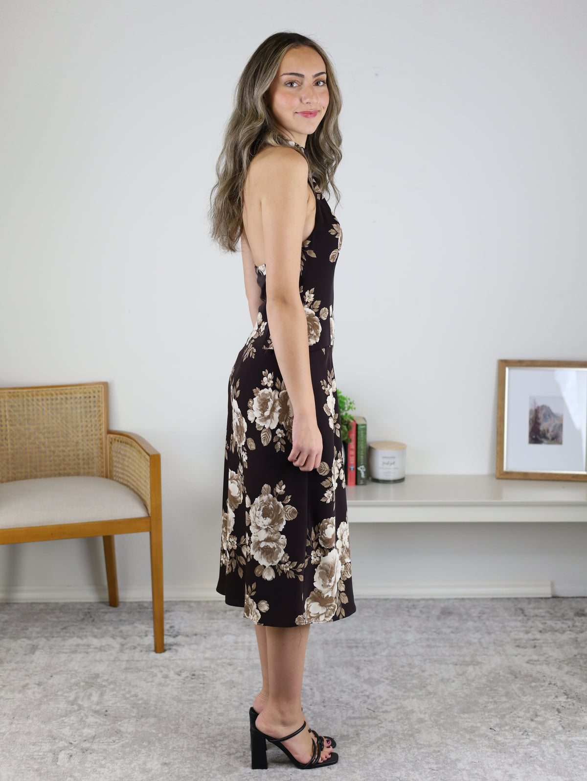 A woman wearing the brown floral Nash Halter Neck Midi Dress with black strappy block-heeled sandals, perfect as a fall wedding guest outfit.