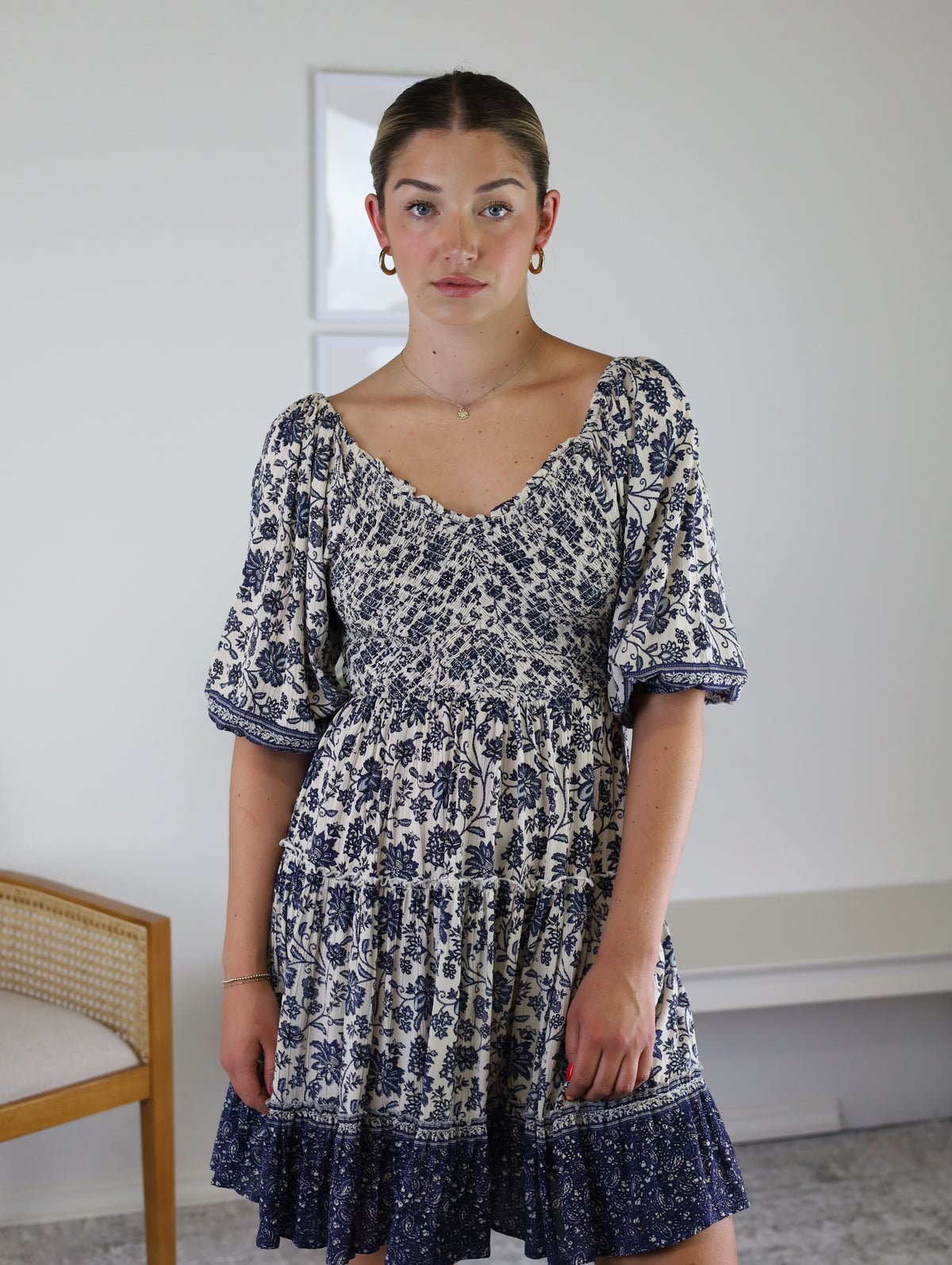 A woman wearing the Leslie Smocked Mini Dress, a cream-colored piece with blue floral patterns, puff sleeves, and a lace-up back.