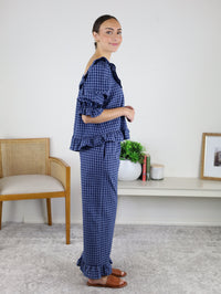 A woman stands indoors wearing the Barron Cotton Gingham Frill Set, a charming blue cotton ensemble featuring a frilled top and matching pants paired with brown sandals. She smiles beside a bench, a low bookshelf filled with books, a plant, and a framed photo—perfectly capturing the comfort and style of women’s loungewear.