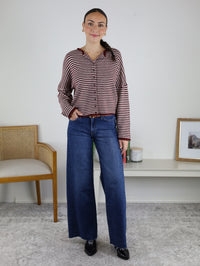 Woman standing indoors wearing the Maren Striped Cardigan, a relaxed fit red and white striped long-sleeve top, paired with blue wide-leg jeans and black shoes. She is smiling with one hand in her pocket. A chair and framed picture are visible in the background.