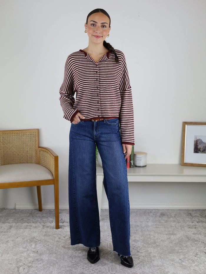 Woman standing indoors wearing the Maren Striped Cardigan, a relaxed fit red and white striped long-sleeve top, paired with blue wide-leg jeans and black shoes. She is smiling with one hand in her pocket. A chair and framed picture are visible in the background.