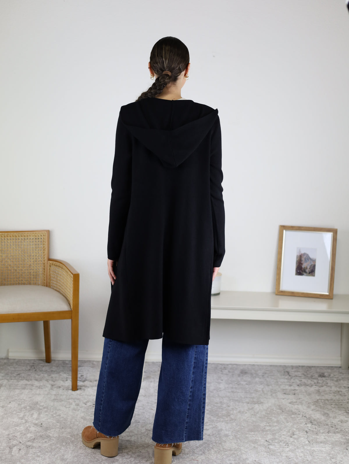 A woman with braided hair, wearing the Pinch Me Long Cardigan styled as a cozy black coat and paired with wide-leg blue jeans, stands facing away in a minimalist room featuring a chair, a framed photo, and a white bench.