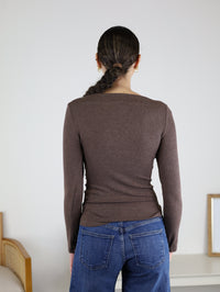 A woman with a long braid, wearing the Tatum Ribbed Boatneck top in brown and blue jeans, stands facing away from the camera in a minimal indoor setting.