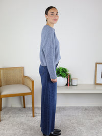 A woman stands sideways in a room, smiling with her hair tied back. She is wearing the Beth Lightweight Cardigan in gray, blue wide-leg jeans, and black shoes. In the background are a chair, shelf, potted plant, and framed picture.