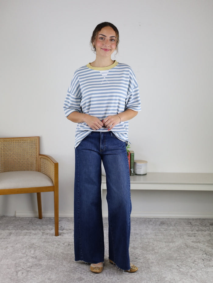 Wearing the Rallie Striped Top in an oversized fit with wide-leg jeans and tan flats, a woman stands indoors, smiling slightly with her hands together in a simply decorated room featuring a chair, shelf, and books.