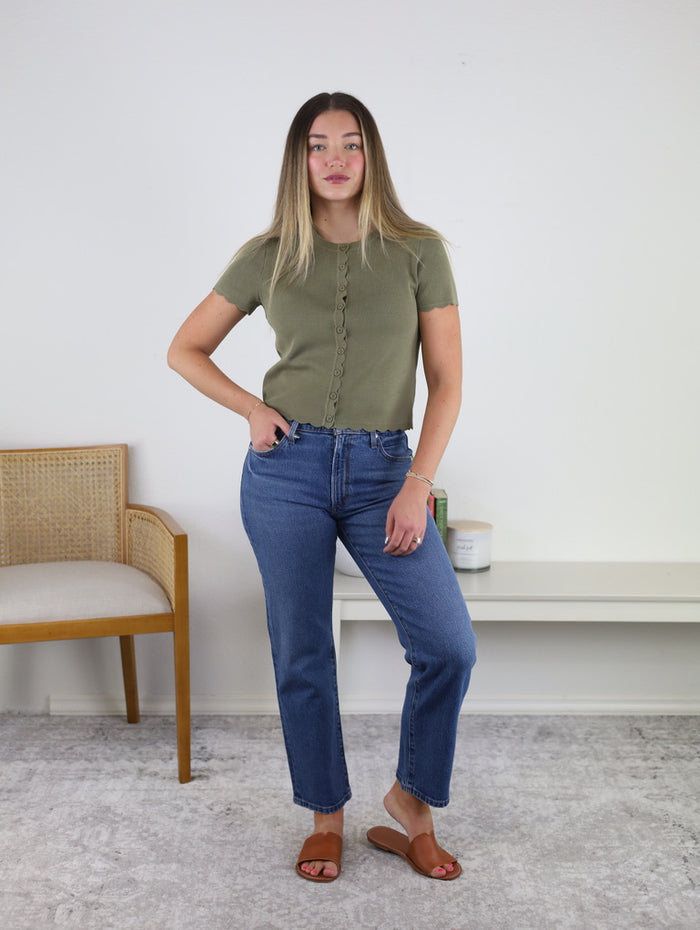 A woman stands indoors on a gray rug, wearing the Aspen Scalloped Cardigan—a green lightweight knit with scalloped trim—paired with blue jeans and brown sandals. Her straight, shoulder-length hair frames her face as she stands near a wooden chair and a white bench.