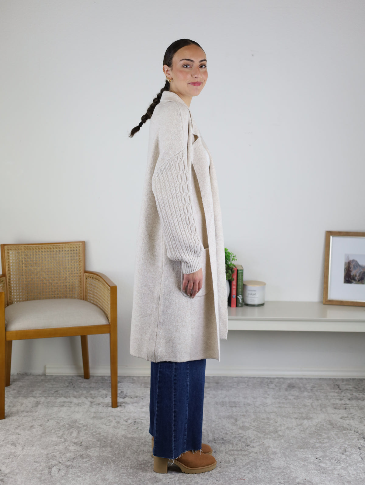 Woman with braided hair stands in profile wearing the Ashford Cable Knit Long Sleeve Knit Coat in beige, styled with blue jeans and tan shoes. She is indoors near a chair and a console table adorned with books, a plant, and framed art.