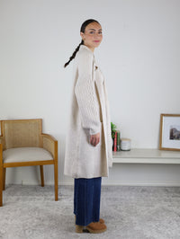 Woman with braided hair stands in profile wearing the Ashford Cable Knit Long Sleeve Knit Coat in beige, styled with blue jeans and tan shoes. She is indoors near a chair and a console table adorned with books, a plant, and framed art.