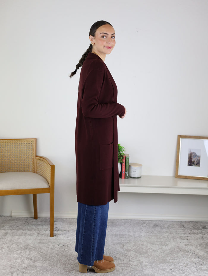 A woman stands sideways indoors, smiling and wearing the Parkland Long Cardigan in maroon, styled with blue jeans and tan shoes. Her hair is braided, and the background features a chair, a bookshelf with books, and a framed picture.