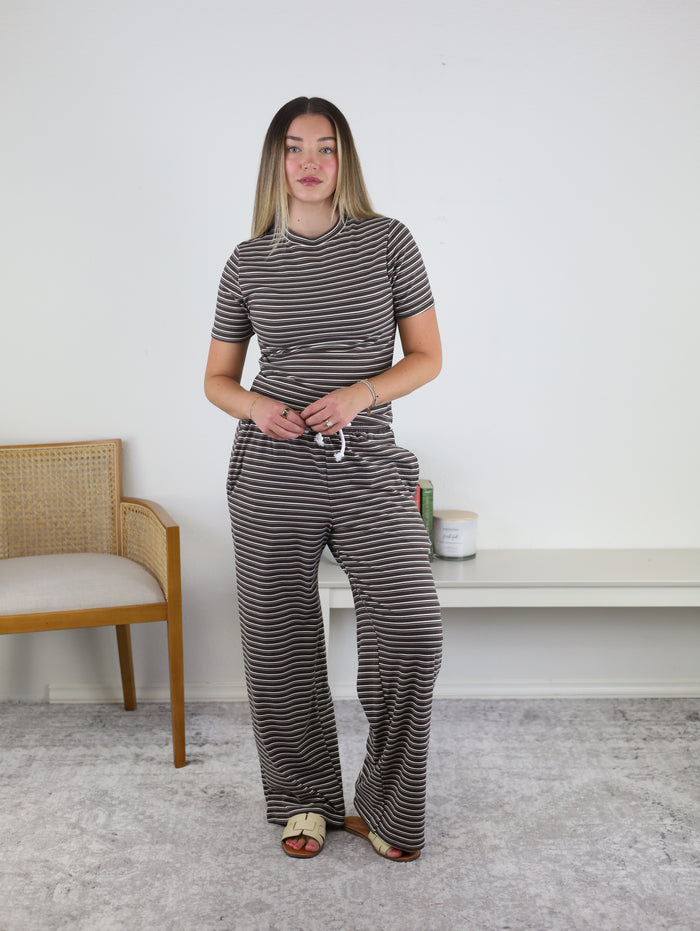 A woman stands indoors wearing the Randi Everyday Striped Lounge Set, which features a soft knit, short-sleeve black and white striped top paired with matching wide-leg pants. She is barefoot, hands clasped together with a neutral expression, with a chair and decor visible in the background.