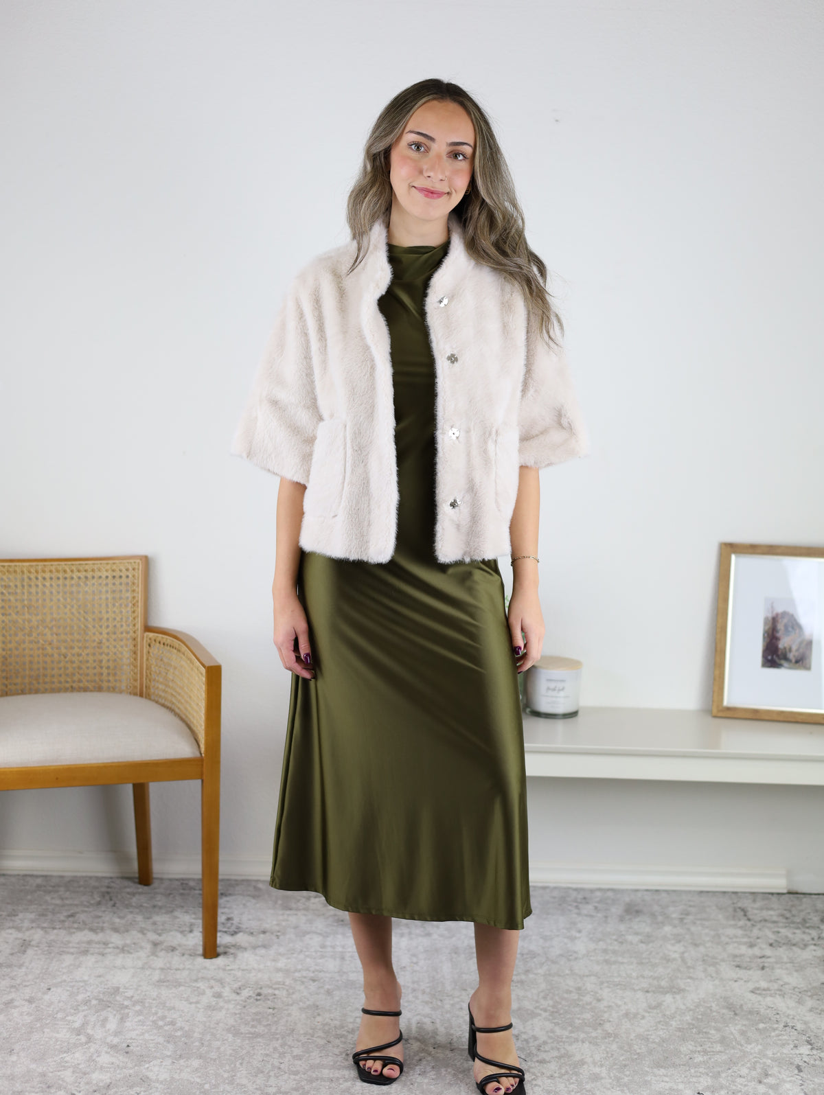 A woman wearing a cream-colored Jordan Fur Cape Sleeve Jacket over a midi-length olive green satin dress.