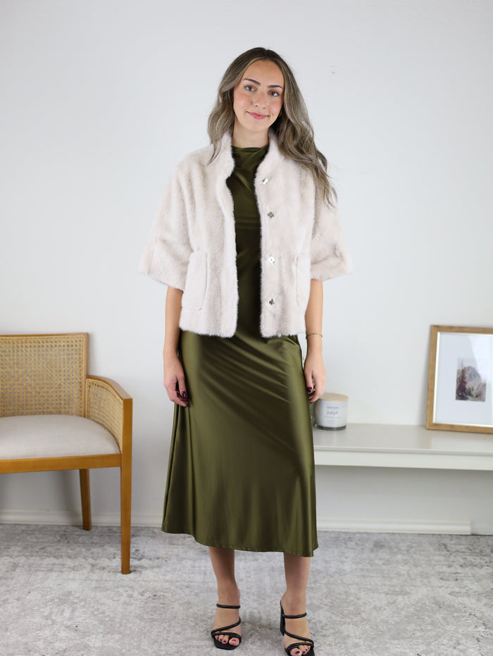 A woman wearing a cream-colored Jordan Fur Cape Sleeve Jacket over a midi-length olive green satin dress.