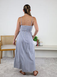 A woman with a braided ponytail is standing indoors with her back to the camera, wearing the Molly Striped Maxi Dress—a blue and white summer cotton maxi dress with adjustable straps—paired with brown sandals. In the background, a chair, books, and a plant are visible.