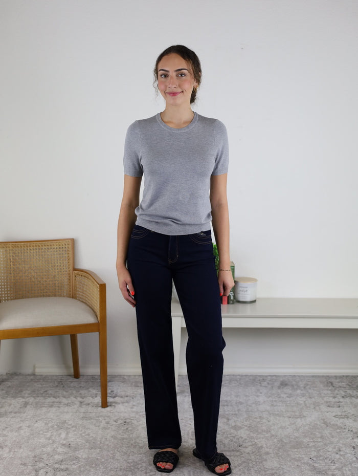 A woman stands indoors on a grey carpet, wearing high-rise Mae Double Pocket Full Length Wide Leg jeans, a grey short-sleeve top, and black sandals. Behind her are a wooden chair with a cushion and a white shelf with decor items.