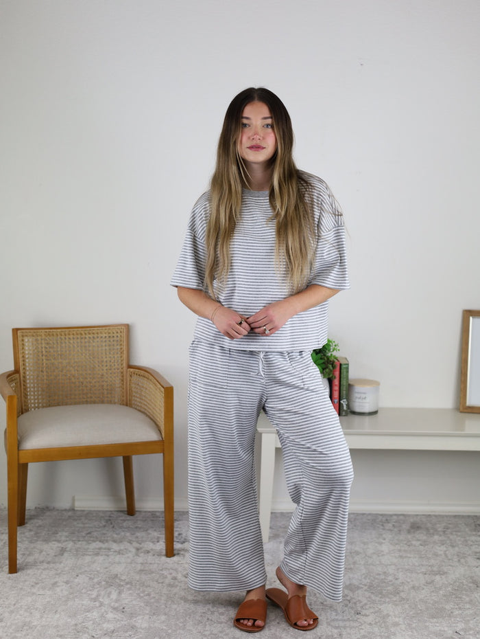 A woman with long hair wears the Alanna Striped Knit Top and Pants Set with sandals, standing indoors by a wooden chair and a white table decorated with decor items.