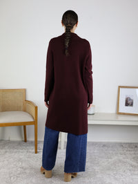 A person with a long braid stands indoors, their back to the camera, wearing the Parkland Long Cardigan in burgundy over blue wide-leg jeans and tan shoes. A chair and a table with a framed photo are in the background, creating a cozy fall outfit scene.