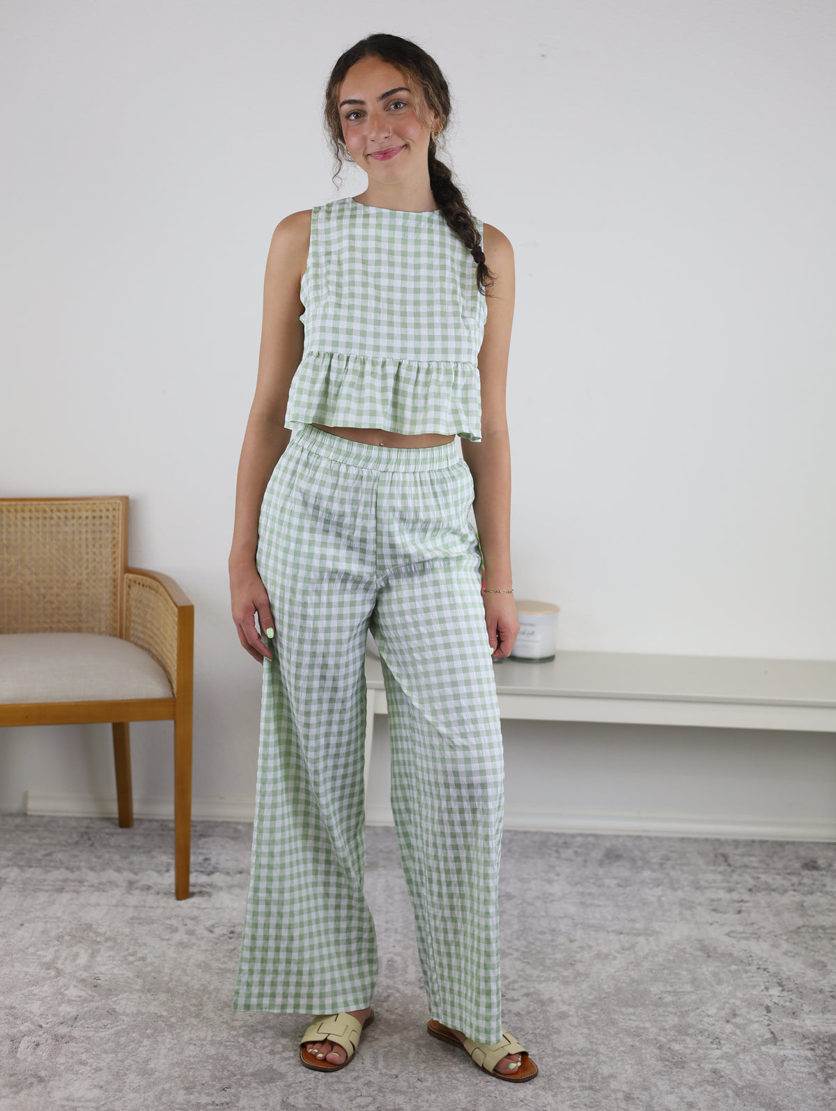 A woman stands indoors wearing the Myra Gingham Print Ruffled Set, which features a sage gingham sleeveless crop top and matching high-waisted wide-leg pants. She has long, braided hair and is smiling. A wooden chair and a bench are visible behind her.