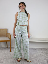 A woman stands indoors wearing the Myra Gingham Print Ruffled Set, which features a sage gingham sleeveless crop top and matching high-waisted wide-leg pants. She has long, braided hair and is smiling. A wooden chair and a bench are visible behind her.
