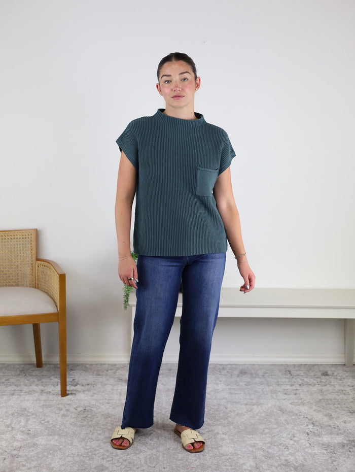 A person stands indoors on a carpet, wearing the Paloma Mock Neck Sweater in green, featuring short sleeves and a front pocket, paired with blue jeans and cream sandals. There is a wooden chair and a white bench in the background.