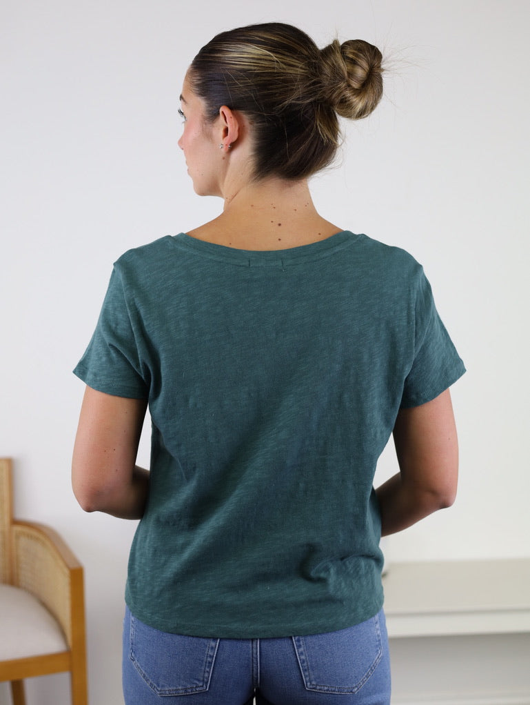 A woman stands facing away, wearing the Nomad V Neck Tee in green and blue jeans. Her hair is tied in a bun. A wooden chair and a white table are in the background against a light wall.