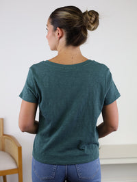 A woman stands facing away, wearing the Nomad V Neck Tee in green and blue jeans. Her hair is tied in a bun. A wooden chair and a white table are in the background against a light wall.