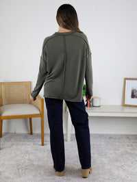 A person with long hair, wearing a green ribbed knit Finley Sweater and navy pants, stands facing away in a minimal room with a chair, shelf, framed photo, and small decor.