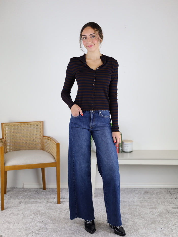 A woman stands indoors, smiling at the camera with one hand in her pocket, wearing the Everette Lightweight Striped Top in black and brown paired with wide-leg blue jeans. In the background, a chair and a white shelf with decor can be seen.