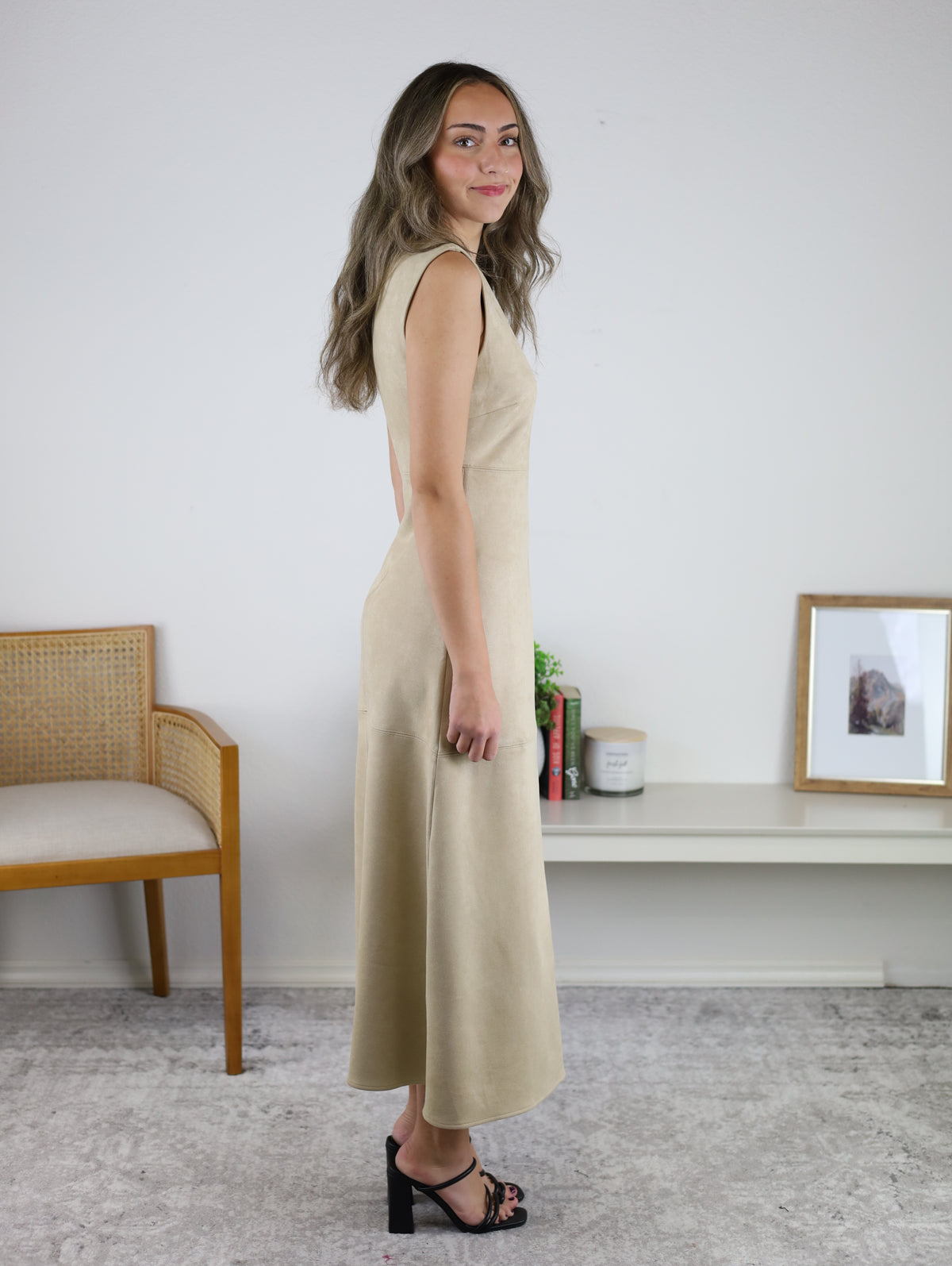 A person standing in profile wearing the tan, sleeveless suede Cole Suede A-Line Maxi Dress and black heeled sandals.