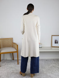 A person with long dark hair in a braid, wearing the elegant Parkland Long Cardigan paired with wide-leg jeans, stands facing away in a minimal room with a chair and framed photo on a bench—a perfect fall outfit.