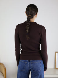 A person with a braided ponytail, wearing the Alexa Snap Button Ribbed Sweater in brown and blue jeans, stands facing away from the camera in a room with neutral decor.