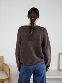 Wearing the Beth Lightweight Cardigan in brown paired with blue jeans, a person with a long braided ponytail stands with their back to the camera in a minimal room featuring a chair and a small table.