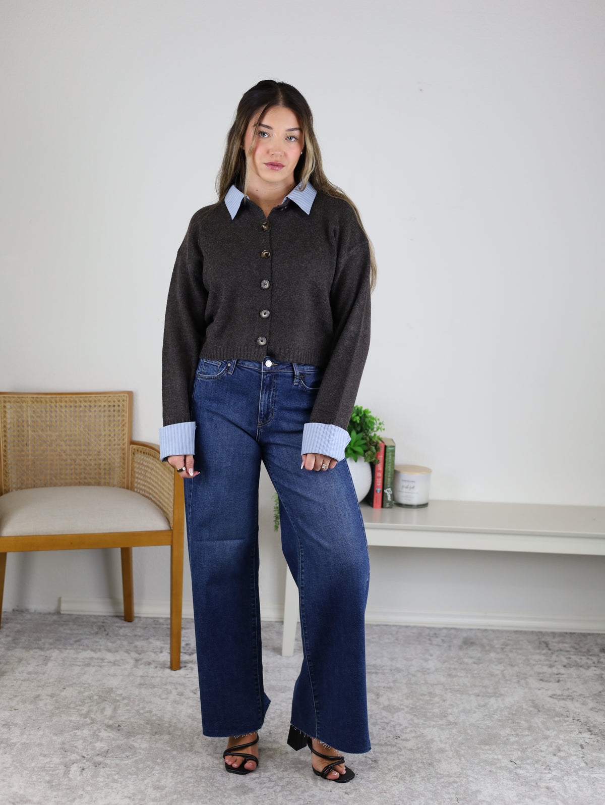 A woman stands indoors wearing the Mani Stripe Trim Button Down Cardigan over a blue collared shirt, wide-leg jeans, and black heels. She poses in front of a white wall, next to a wooden chair and a table with books, a candle, and a plant.