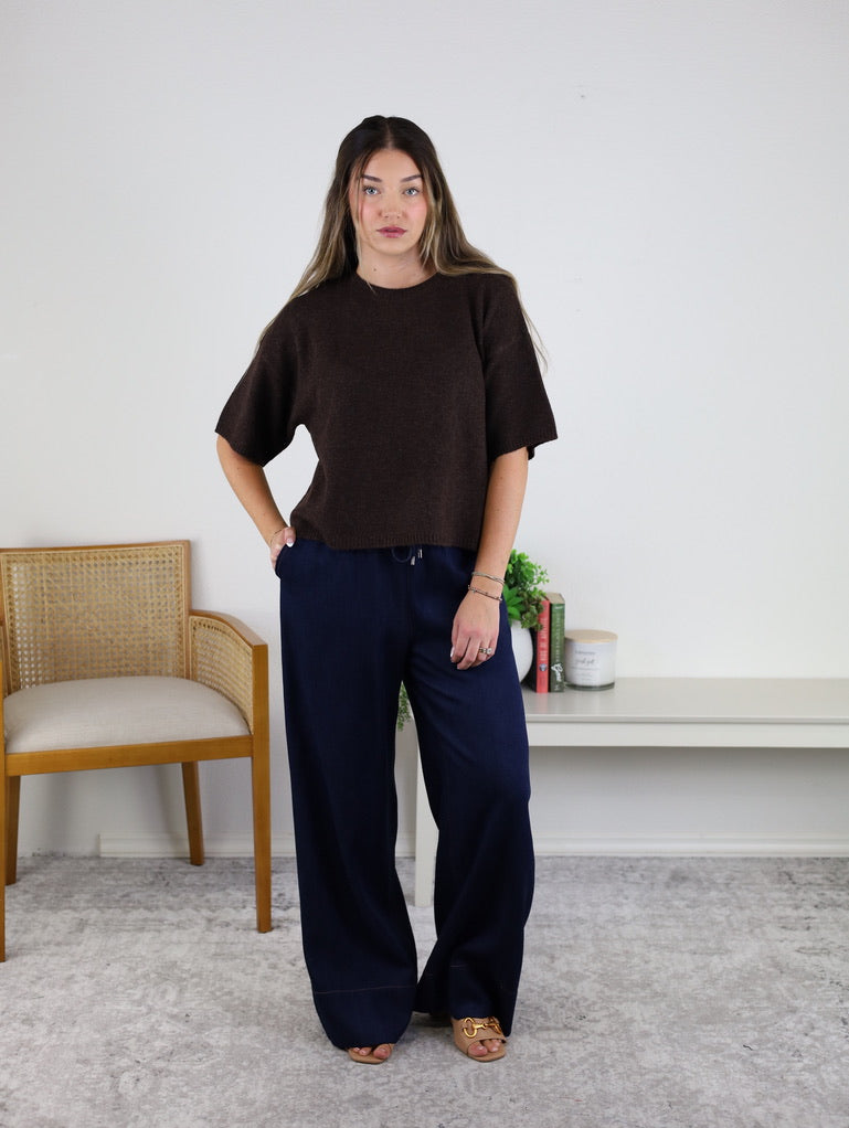 A woman with long hair stands indoors, wearing the West Raw Edge Sweater—a relaxed brown cotton-acrylic pullover—paired with navy wide-leg pants and sandals. She poses with one hand on her hip beside a wooden chair, a small white shelf, books, and plants.