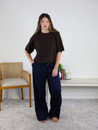 A woman with long hair stands indoors, wearing the West Raw Edge Sweater—a relaxed brown cotton-acrylic pullover—paired with navy wide-leg pants and sandals. She poses with one hand on her hip beside a wooden chair, a small white shelf, books, and plants.