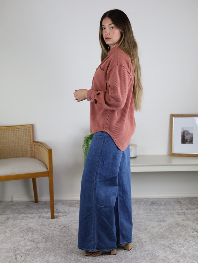 A woman with long hair wears the Larkin Rib Knit Button Up in rust and wide-leg blue jeans indoors, with a chair, plant, and shelf with framed art in the background.