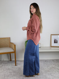 A woman with long hair wears the Larkin Rib Knit Button Up in rust and wide-leg blue jeans indoors, with a chair, plant, and shelf with framed art in the background.