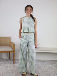 A woman stands indoors wearing the Myra Gingham Print Ruffled Set, which features a sleeveless sage gingham peplum top with ruffle details and matching high-waisted wide-leg pants. She smiles with one hand in her pocket, while a chair and white bench are visible in the background.