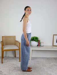 A woman stands in profile indoors, wearing a white tank top, brown shoes, and the Marigold Acid Wash Wide Leg Denim. Behind her are a wooden chair, a low table with books, a plant, and a framed picture against a white wall.