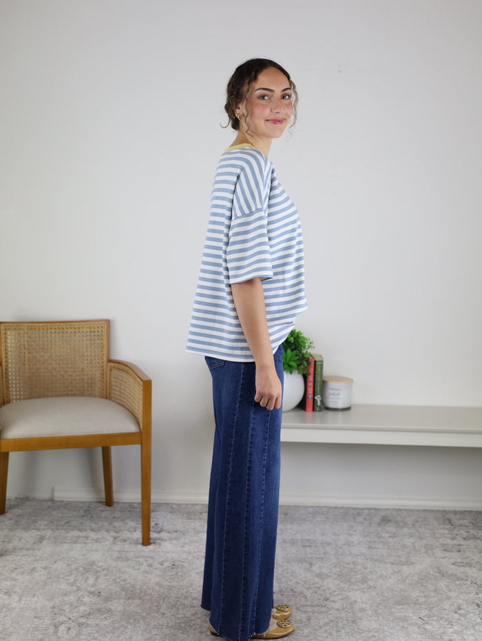 A woman stands sideways, smiling, wearing the Rallie Striped Top—an oversized fit blue and white striped shirt—paired with wide-leg jeans. She is indoors, near a wooden chair and a white shelf adorned with books, a candle, and a small plant.